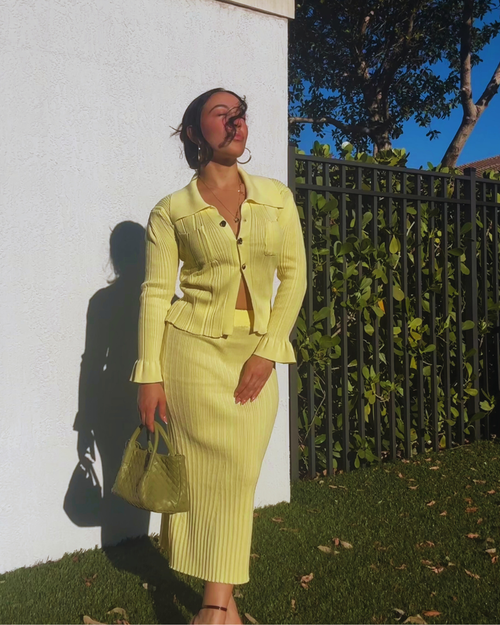 Woman in a yellow outfit standing outdoors with a white wall and greenery in the background